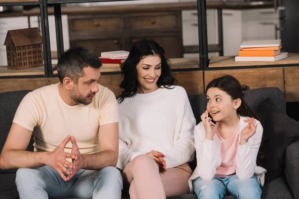 Niño feliz hablando en teléfono inteligente cerca de los padres en la sala de estar - foto de stock