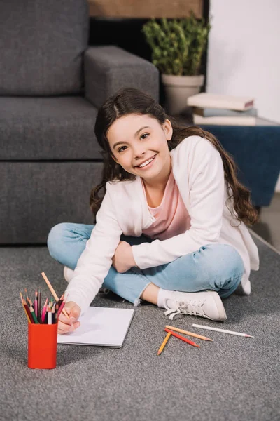 Enfant souriant regardant la caméra tout en tenant un crayon près du papier dans le salon — Photo de stock
