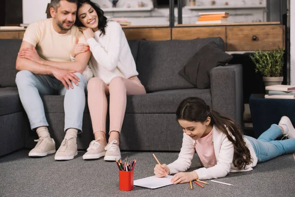 Selektiver Fokus des niedlichen Kindes, das auf dem Boden liegt und in der Nähe glücklicher Eltern auf Papier zeichnet — Stockfoto