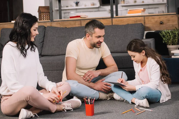 Glückliche Eltern sitzen auf dem Boden neben niedlichen Tochter Zeichnung auf Papier — Stockfoto