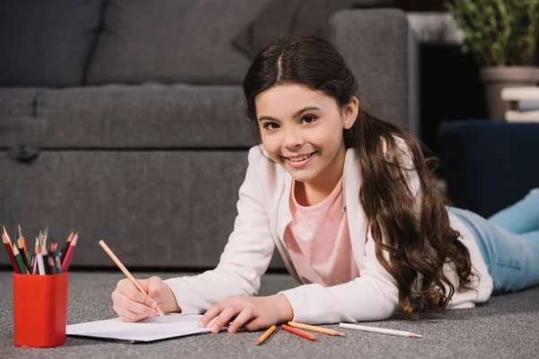 Cheerful kid lying on floor and drawing on paper in living room — Stock Photo