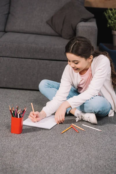 Niño alegre sentado en el suelo y dibujo sobre papel en la sala de estar - foto de stock