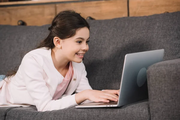 Enfant heureux couché sur le canapé et en utilisant un ordinateur portable dans le salon — Photo de stock