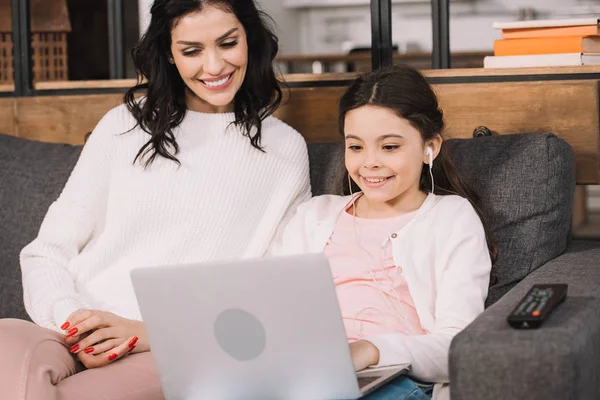 Fröhliche Mutter blickt auf Laptop in der Nähe Tochter hört Musik in Kopfhörer — Stockfoto