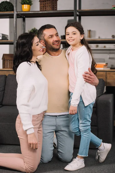 Alegre hombre abrazando atractiva esposa y linda hija en casa - foto de stock