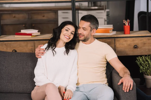 Feliz hombre abrazando alegre esposa mientras está sentado en sofá en casa - foto de stock