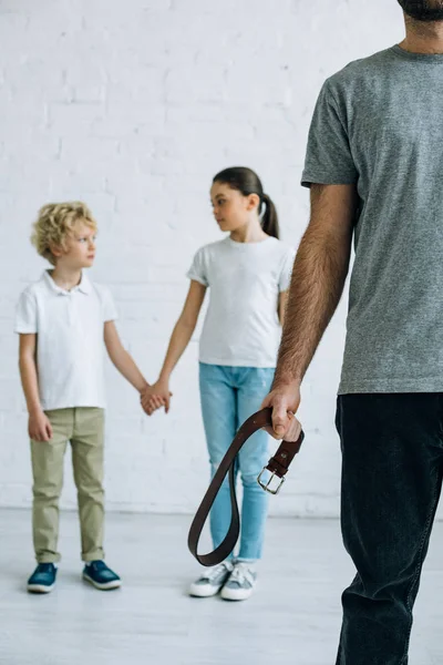 Partial view of abusive father with belt and kids holding hands — Stock Photo