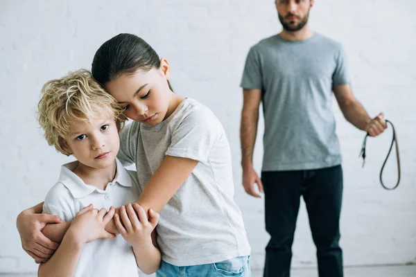 Partial view of abusive father with belt and embracing kids at home — Stock Photo