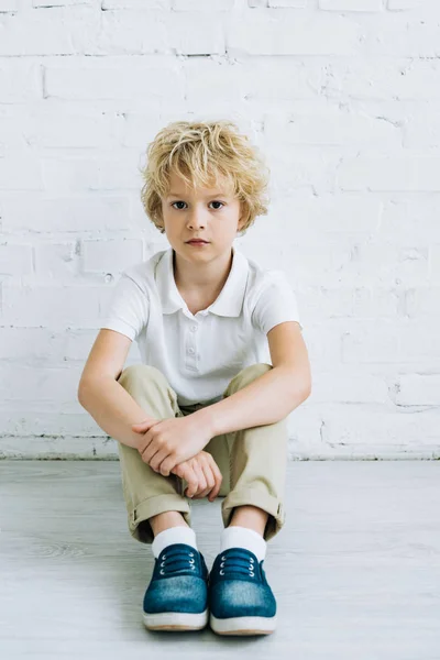 Niño preadolescente molesto sentado en el suelo y mirando a la cámara en casa - foto de stock