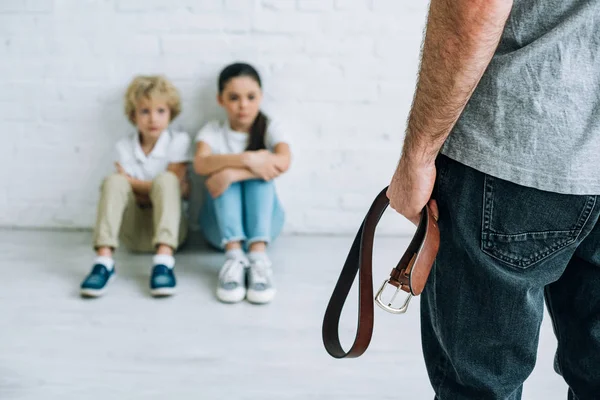 Vista recortada de un padre abusivo sujetando el cinturón y niños tristes sentados en el suelo - foto de stock