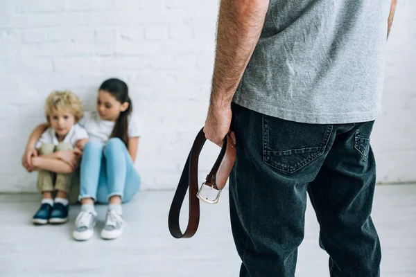 Vista recortada de un padre abusivo sujetando el cinturón y niños tristes sentados en el suelo - foto de stock