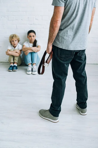 Ausgeschnittener Blick auf missbräuchlichen Vater mit Gürtel und traurige Kinder auf dem Boden — Stockfoto