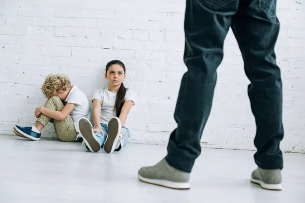 Vista recortada de un padre abusivo sujetando el cinturón y niños tristes sentados en el suelo - foto de stock