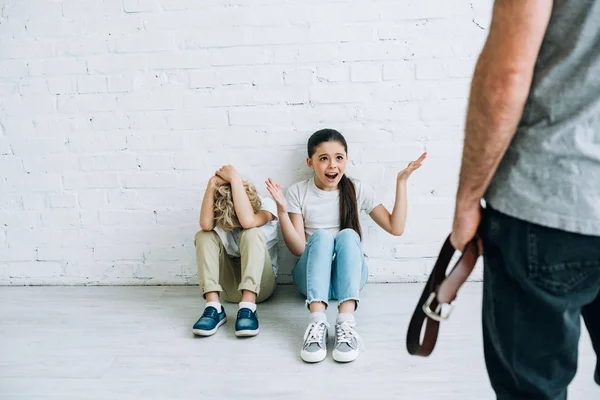 Cropped view of abusive father holding belt and scared kids sitting on floor — Stock Photo