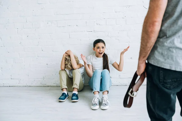 Vista recortada de un padre abusivo sujetando el cinturón y niños asustados sentados en el suelo - foto de stock