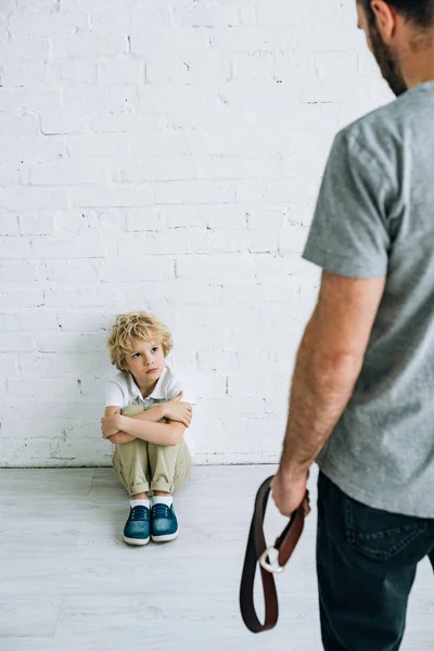 Cropped view of father holding belt and sad son sitting on floor — Stock Photo