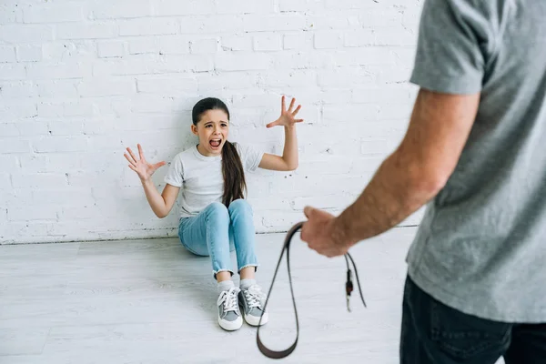 Cropped view of father holding belt and scared daughter sitting on floor — Stock Photo