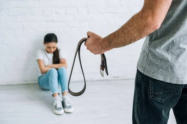 Cropped view of father holding belt and sad daughter sitting on floor — Stock Photo