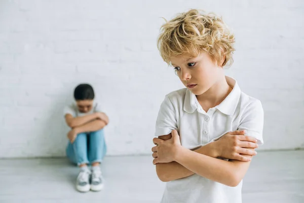 Triste chico de pie con los brazos cruzados mientras hermana sentada en el suelo y llorando en casa - foto de stock