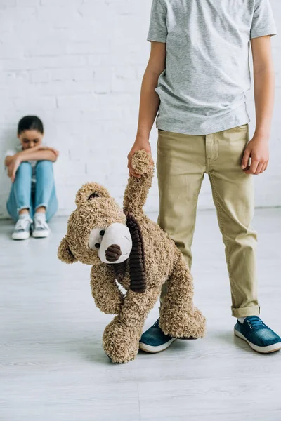 Recortado vista de preadolescente niño sosteniendo osito de peluche mientras su hermana llorando en casa - foto de stock