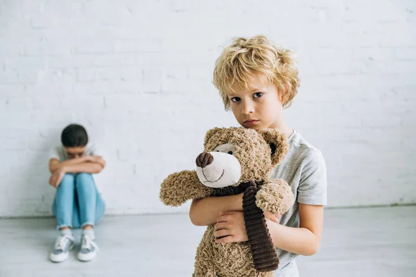 Хлопчик тримає плюшевого ведмедя, а його сестра плаче вдома — стокове фото