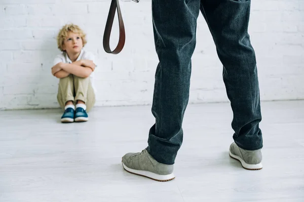 Partial view of abusive father holding belt and son sitting on floor — Stock Photo