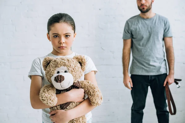 Partial view of abusive father with belt and sad daughter holding teddy bear — Stock Photo