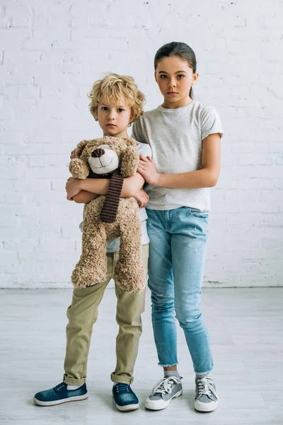 Full length view of sad sister and brother with teddy bear at home — Stock Photo