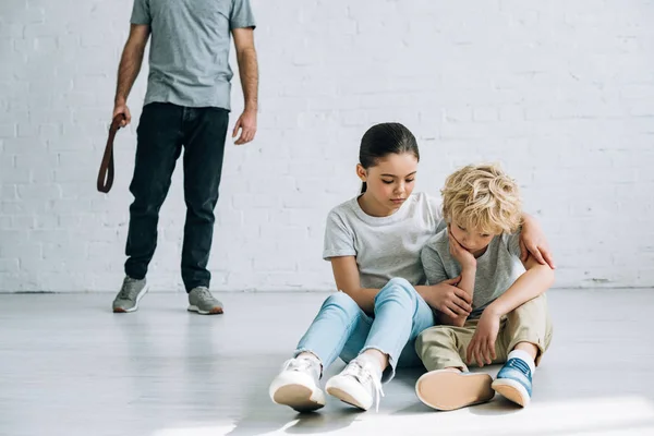 Partial view of abusive father and sad kids sitting on floor — Stock Photo