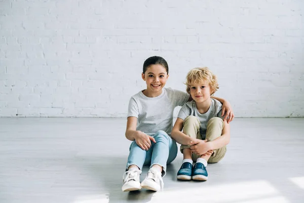 Lächelnde Schwester umarmt Bruder, während sie auf dem Boden sitzt — Stockfoto