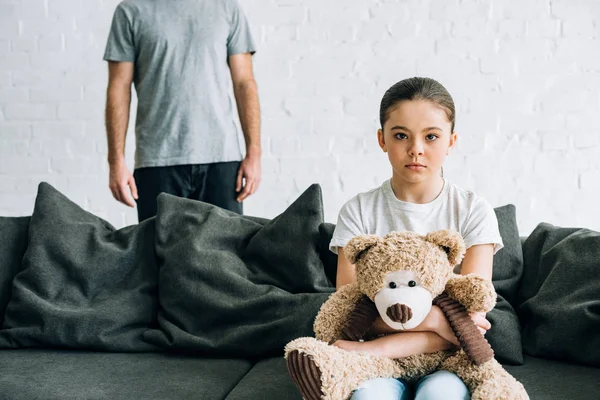 Vista parcial del padre y la triste hija con oso de peluche sentado en el sofá - foto de stock