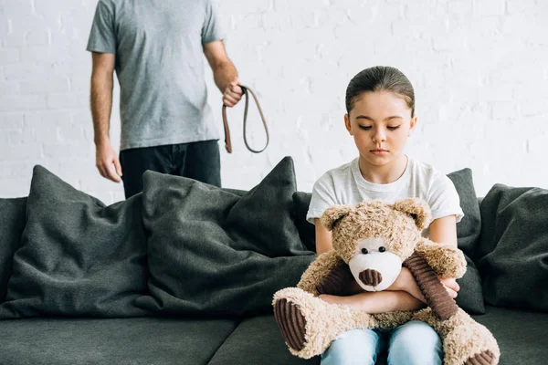 Vista parcial del padre abusivo con cinturón e hija triste con oso de peluche sentado en el sofá - foto de stock