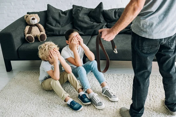 Partial view of father with belt and upset kids sitting on floor — Stock Photo