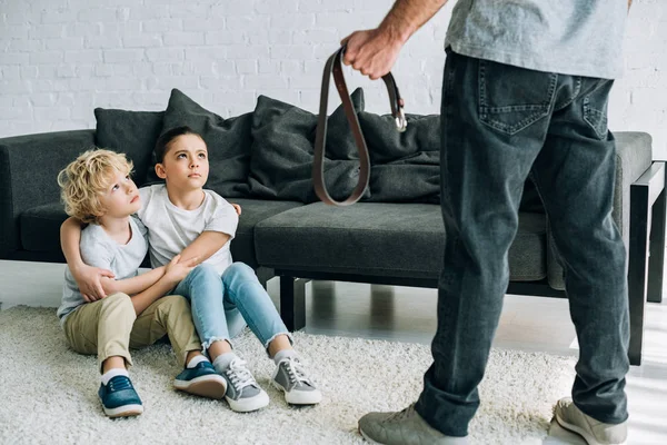 Partial view of father with belt and upset kids sitting on floor — Stock Photo