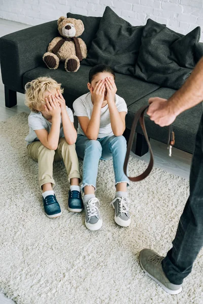 Visão parcial do pai com cinto e crianças chateadas sentadas no chão — Fotografia de Stock