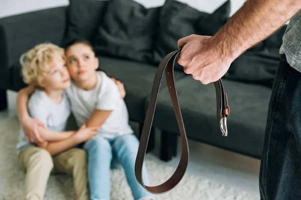 Partial view of father with belt and upset kids sitting on floor — Stock Photo