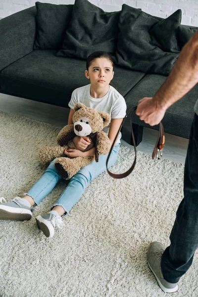 Overhead view of abusive father holding belt and scared daughter with teddy bear sitting on carpet — Stock Photo