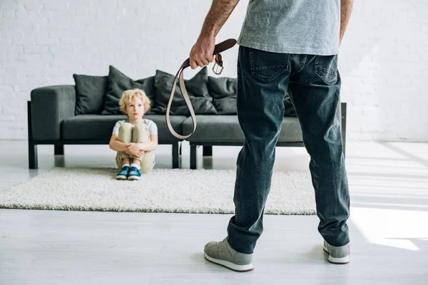 Partial view of abusive father holding belt and sad son sitting on carpet — Stock Photo
