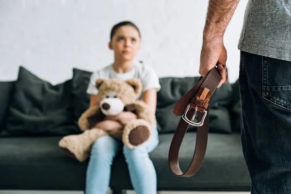 Partial view of abusive father holding belt and scared daughter with teddy bear sitting on sofa — Stock Photo