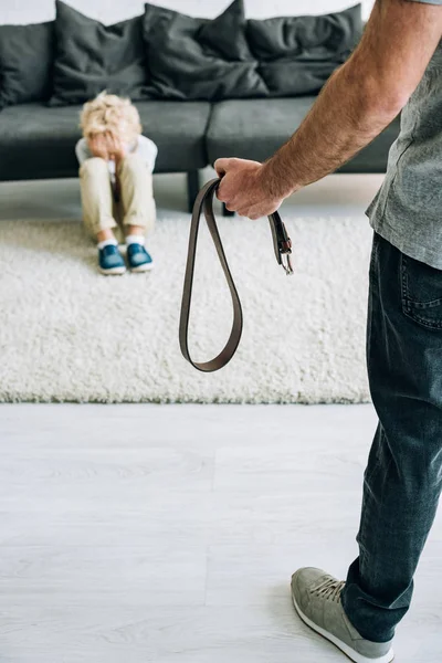 Partial view of abusive father holding belt and sad son sitting on carpet — Stock Photo