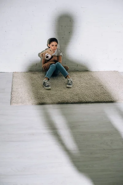 Enfant effrayé avec ours en peluche assis sur le tapis et grande silhouette — Photo de stock