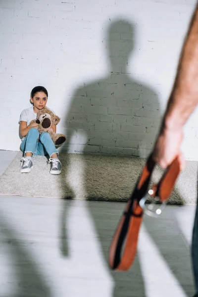 Обрезанный вид отца, держащего ремень, и испуганная дочь, сидящая на ковре рядом с его силуэтом — стоковое фото