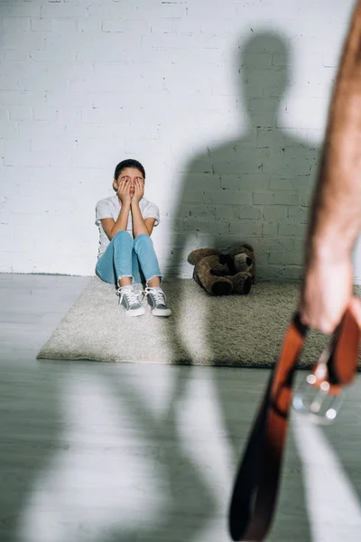 Cropped view of father holding belt and scared daughter sitting on carpet near his silhouette — Stock Photo