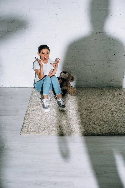 Big silhouette and scared kid with teddy bear screaming while sitting on carpet — Stock Photo