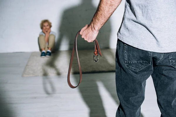 Cropped view of father holding belt and scared son sitting on carpet near his silhouette — Stock Photo