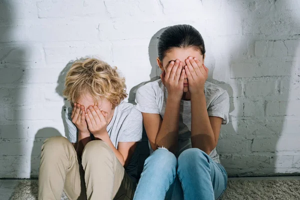 Niños asustados cubriendo los ojos con las manos mientras están sentados en la alfombra - foto de stock