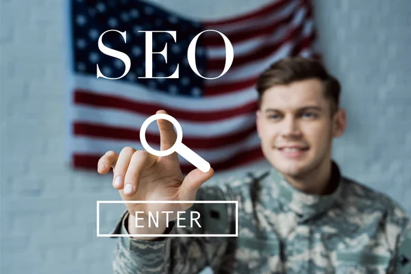 Selektiver Fokus eines glücklichen Mannes in Militäruniform, der mit dem Finger auf den SEO-Schriftzug zeigt — Stockfoto