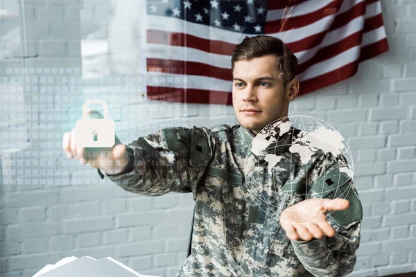 Hombre guapo en uniforme de camuflaje apuntando con el dedo al candado virtual cerca de la bandera americana - foto de stock