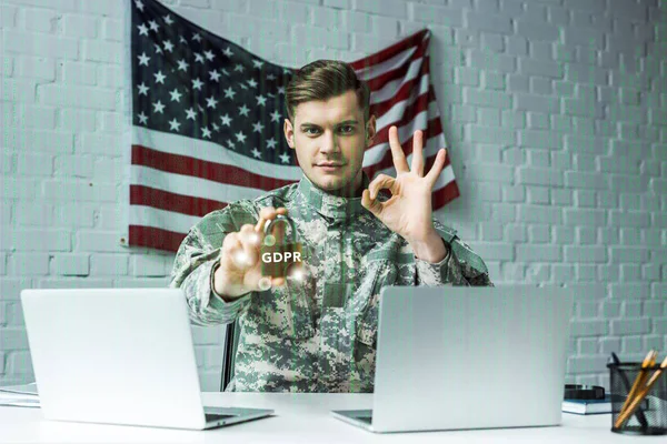 Uomo in uniforme militare in possesso di lucchetto con scritte gdpr vicino computer portatili e mostrando segno ok — Foto stock