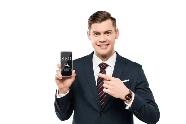 Homem de negócios feliz em terno apontando com o dedo para o smartphone com seo lettering na tela isolada no branco — Fotografia de Stock